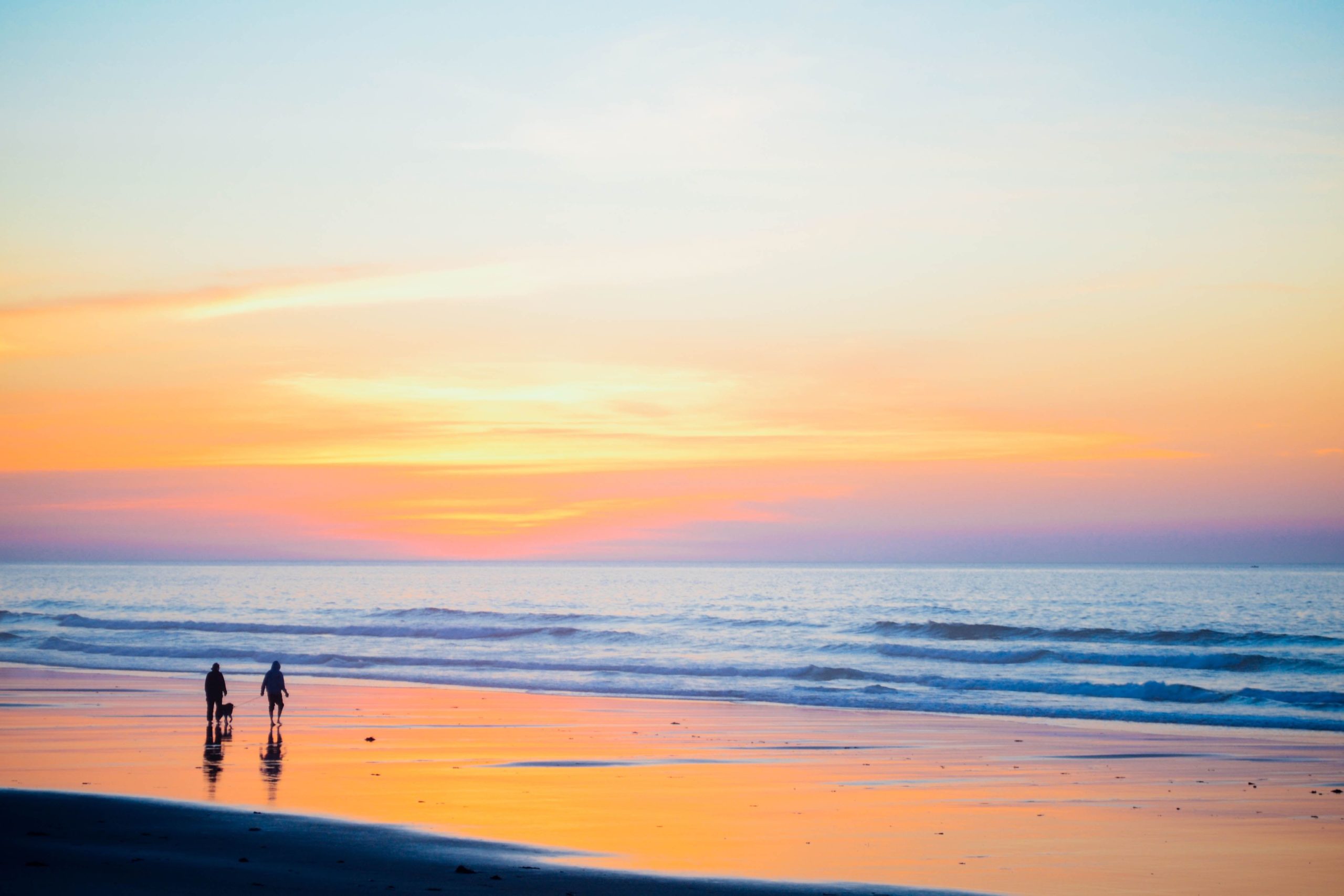 Zwei Menschen gehen bei Sonnenuntergang an einem Sandstrand entlang, mit ruhigen Wellen und einem farbenfrohen Himmel, der sich im nassen Ufer spiegelt.