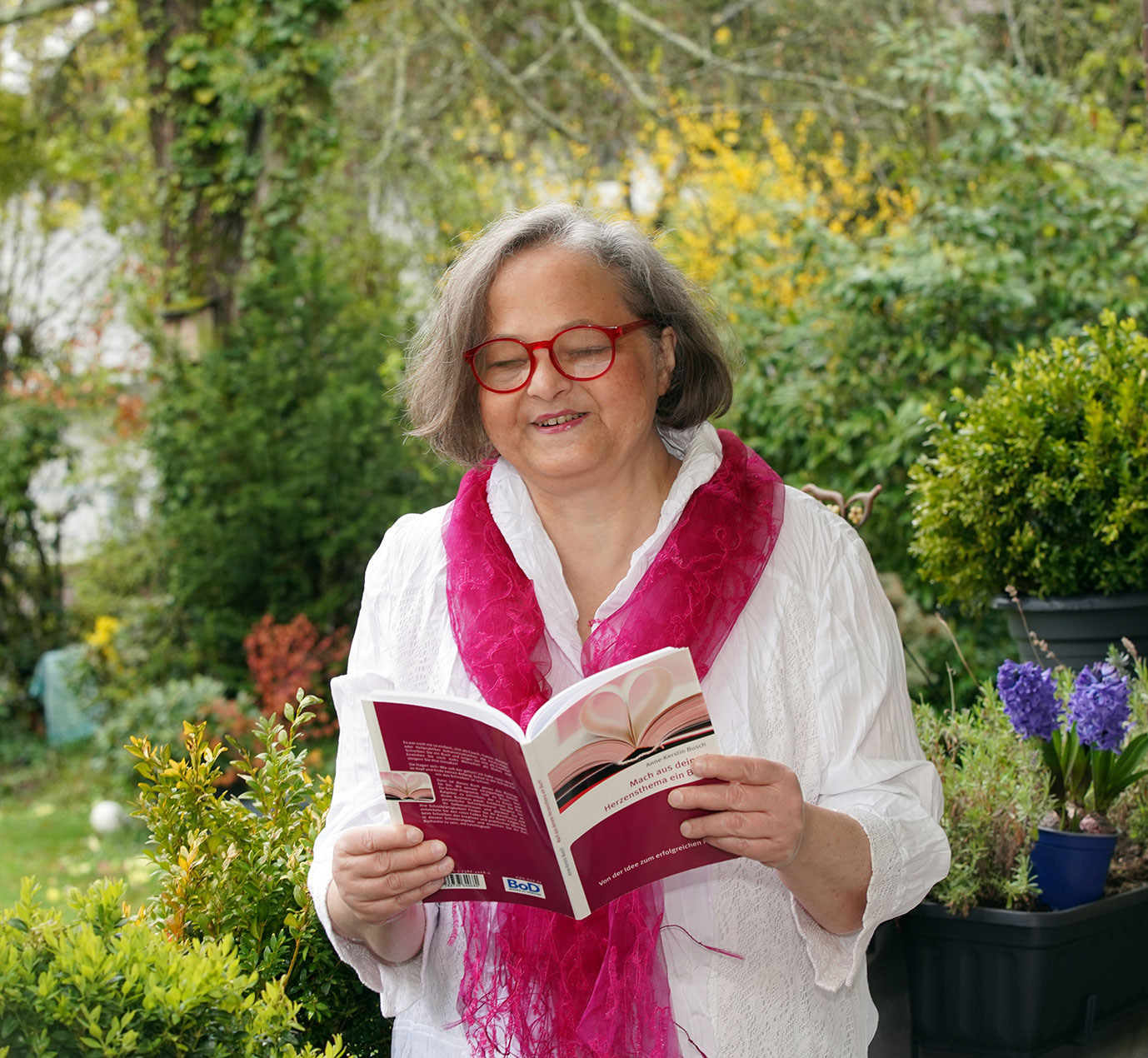 Anne-Kerstin Busch mit ihrem Buch "Mach aus deinem Herzensthema ein Buch. Grüne Sträucher und Blumen im Hintergrund 